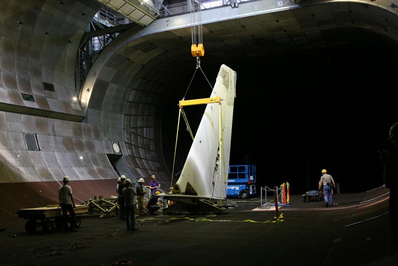 A 40-ton crane carefully lowers an aircraft tail from the rafters down through the open doors of the wind tunnel's roof. Credits - NASA / Eric James.