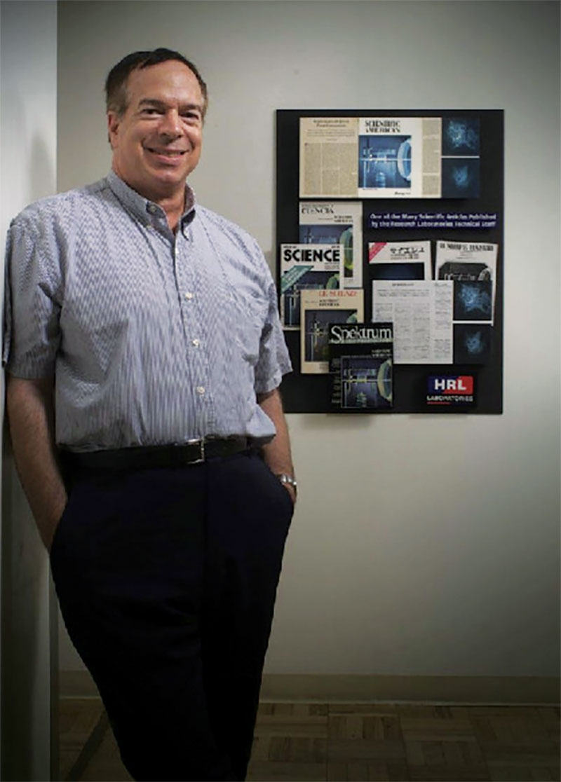 David Pepper next to a poster at HRL of his January 1986 article in Scientific American, “Applications of Optical Phase Conjugation,” translated into various languages. Photo courtesy of Pepper.