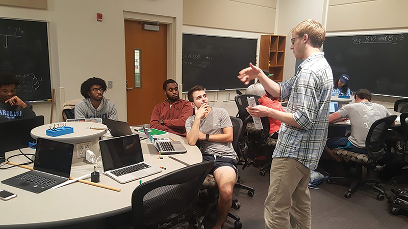 Teaching assistant Ben Kaiser (standing) discusses a torque activity with a student group during an introductory physics course at University of North Carolina at Chapel Hill. Photo by Shane Brogan.