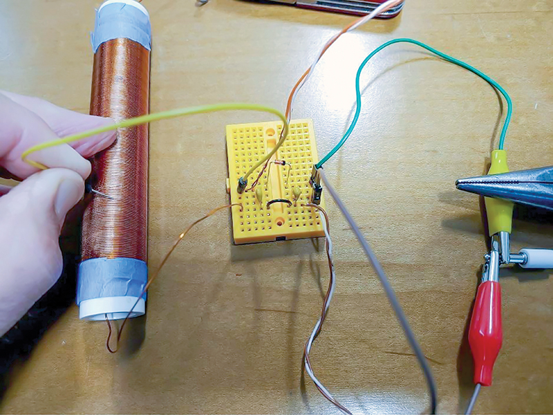 A working radio setup. A germanium (Ge) diode spans the “trough” that runs down the middle of the breadboard circuit and two capacitors on either side of the diode. An inductor tuning coil is on the left. The yellow wire runs from the Ge diode to the tuning coil; sliding it along the coil changes the received station. The breadboard-to-alligator clips (green and brown wires) are clipped to 2/3 connections of the audio jack attached to earbuds. The antenna and the ground are the orange-and-white and brown-and-white wires, respectively.