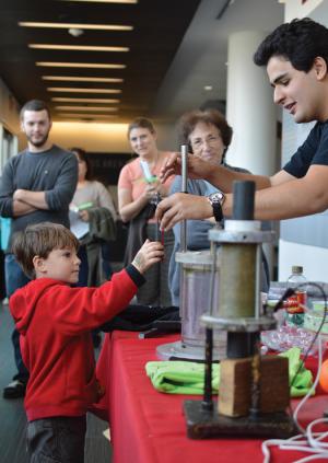 Many SPS and Sigma Pi Sigma chapters rely on both internal and external funding sources  to bring science outreach activities to the public. Pictured here, a crowd gathers around the SPS table during Family Weekend at Rensselaer Polytechnic  Institute. Josue San Emeterio, the student team leader, explains the chaotic double pendulum to a young scientist. Photo by Kelsie Larson.